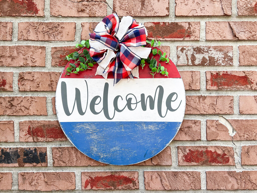 Red white and blue Welcome round pine door hanger with plaid burlap bow and eucalyptus greenery hung on red brick wall hero shot close up