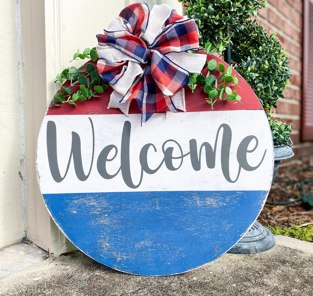 Red white and blue Welcome round pine door hanger with plaid burlap bow and eucalyptus greenery leaning against white door frame with boxwood topiary on brick porch product hero shot