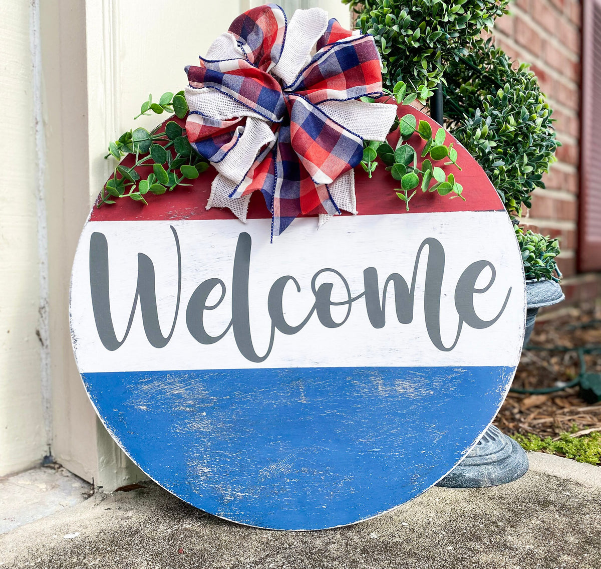 Red white and blue Welcome round pine door hanger with plaid burlap bow and eucalyptus greenery leaning against white door frame with boxwood topiary on brick porch product hero shot