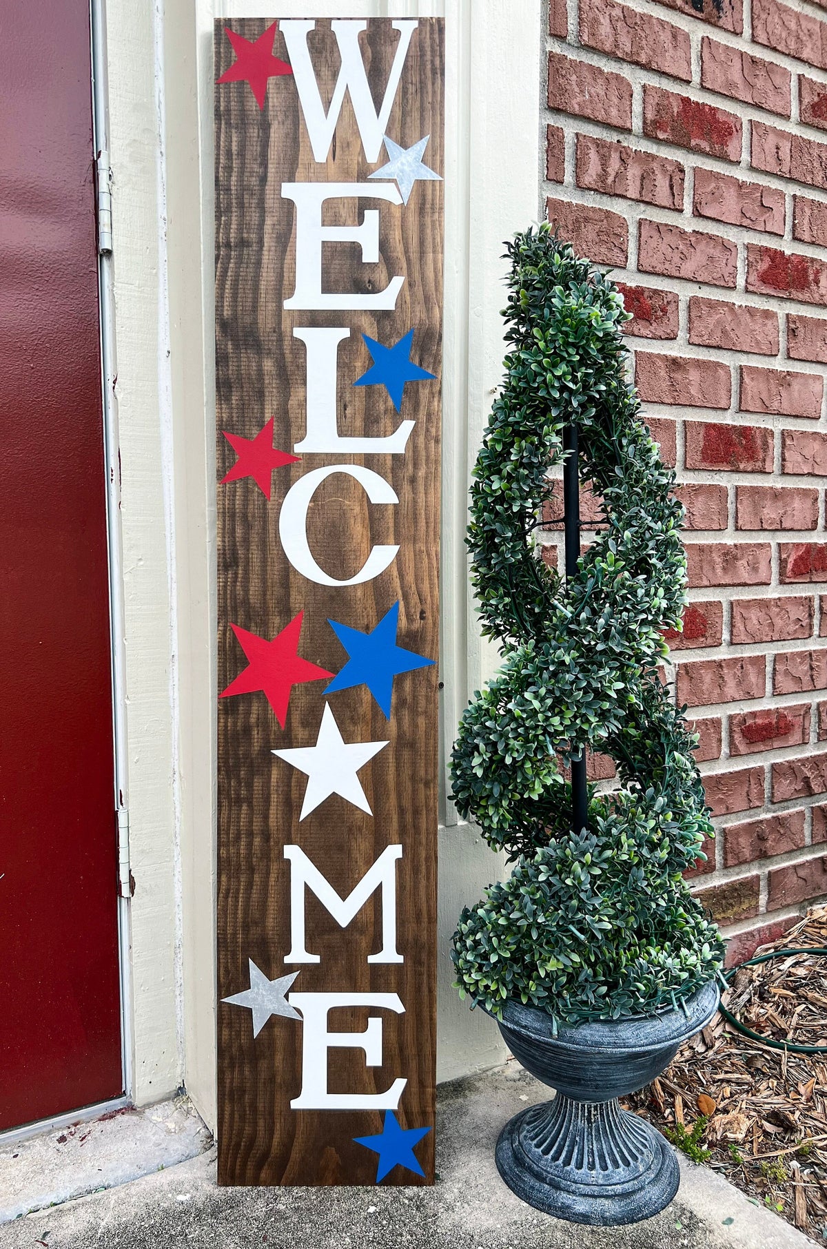 Handpainted Welcome porch leaner sign with red white and blue stars on dark walnut stained white pine board leaning against white door frame close up