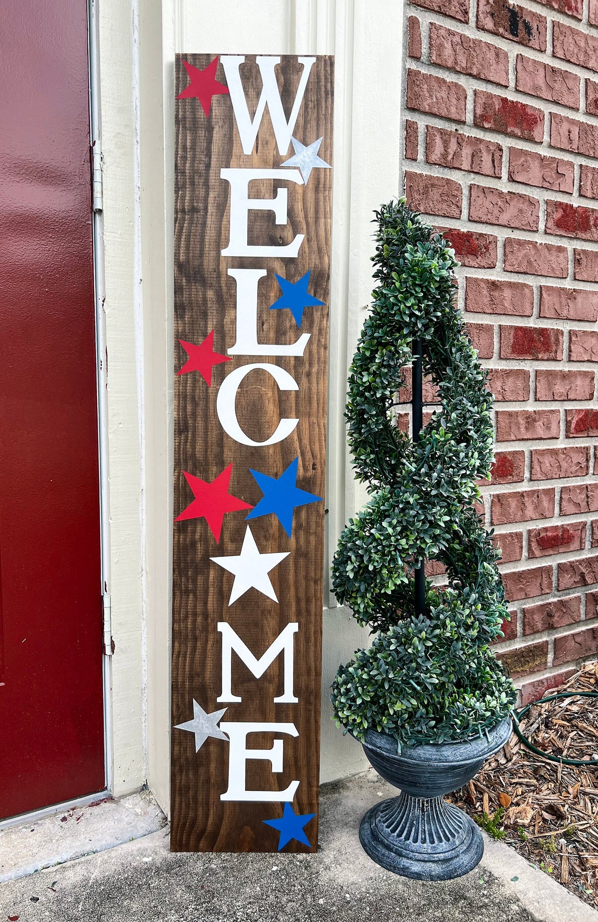 Handpainted Welcome porch leaner sign with red white and blue stars on dark walnut stained white pine board leaning against red door close up