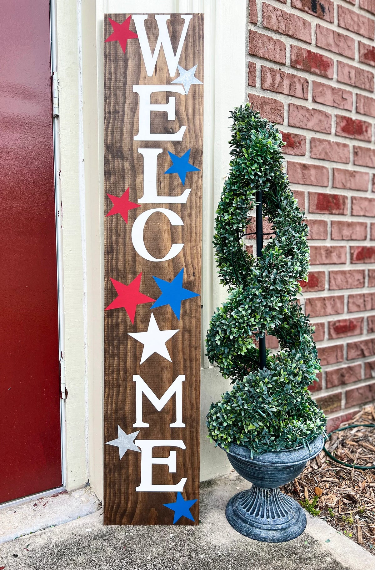 Handpainted Welcome porch leaner sign with red white and blue stars on dark walnut stained white pine board leaning against white door frame with boxwood topiary