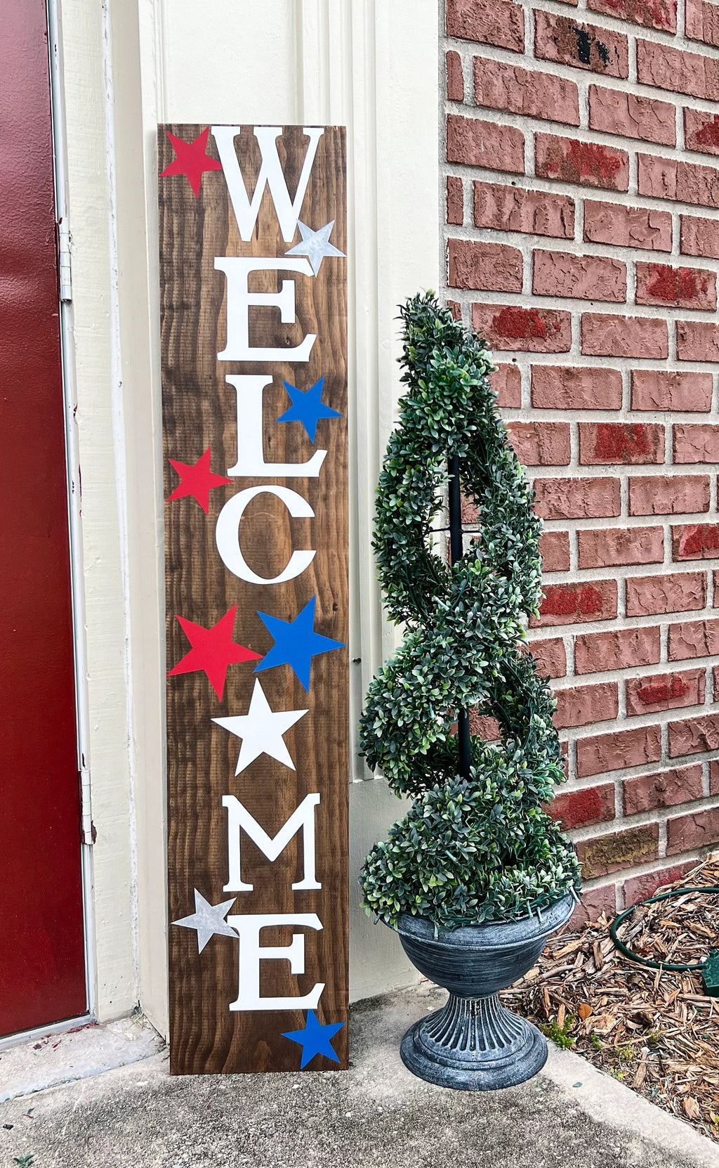 Handpainted Welcome porch leaner sign with red white and blue stars on dark walnut stained white pine board leaning against red door with boxwood topiary