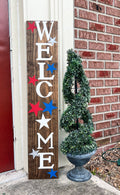 Handpainted Welcome porch leaner sign with red white and blue stars on dark walnut stained white pine board leaning against red door with boxwood topiary