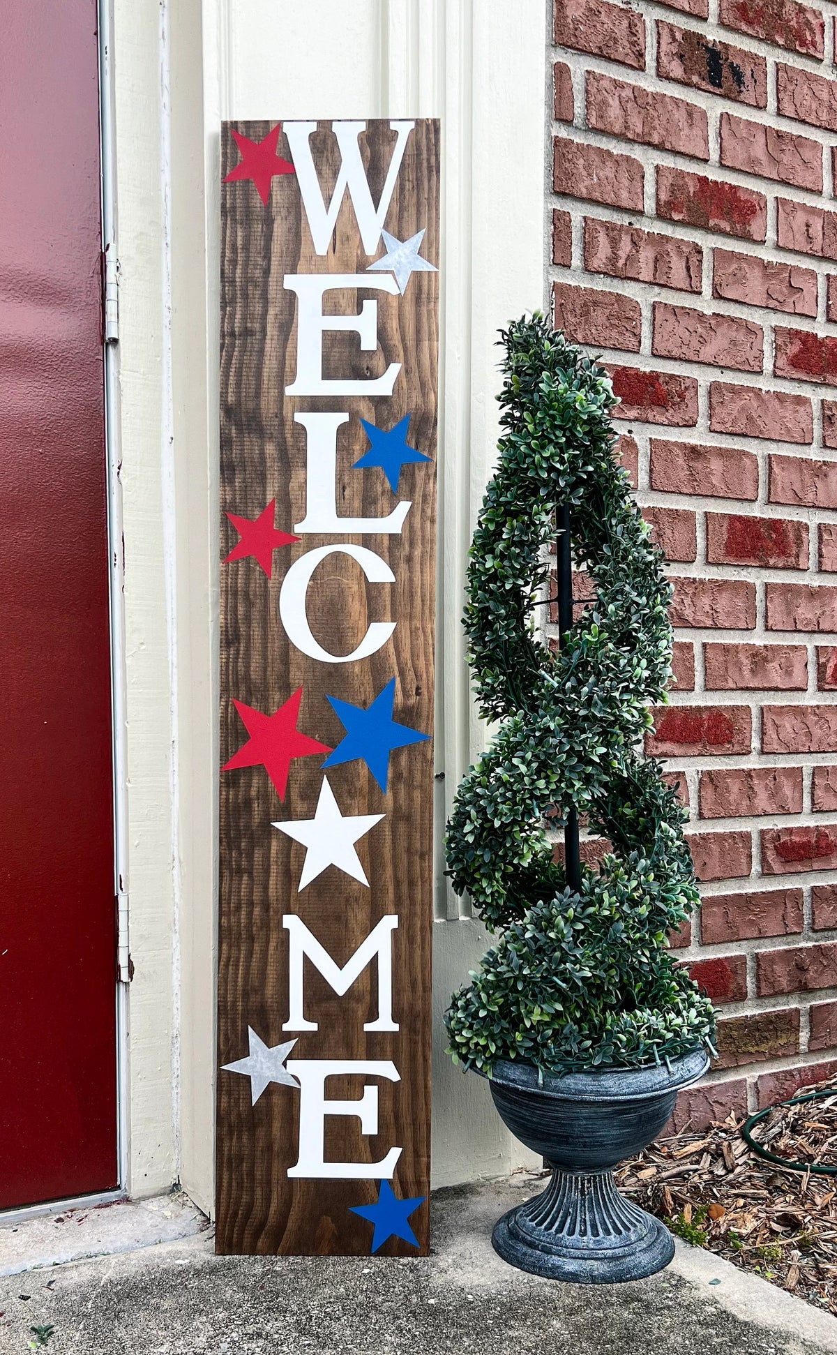 Handpainted Welcome porch leaner sign with red white and blue stars on dark walnut stained white pine board leaning against white door frame wide shot