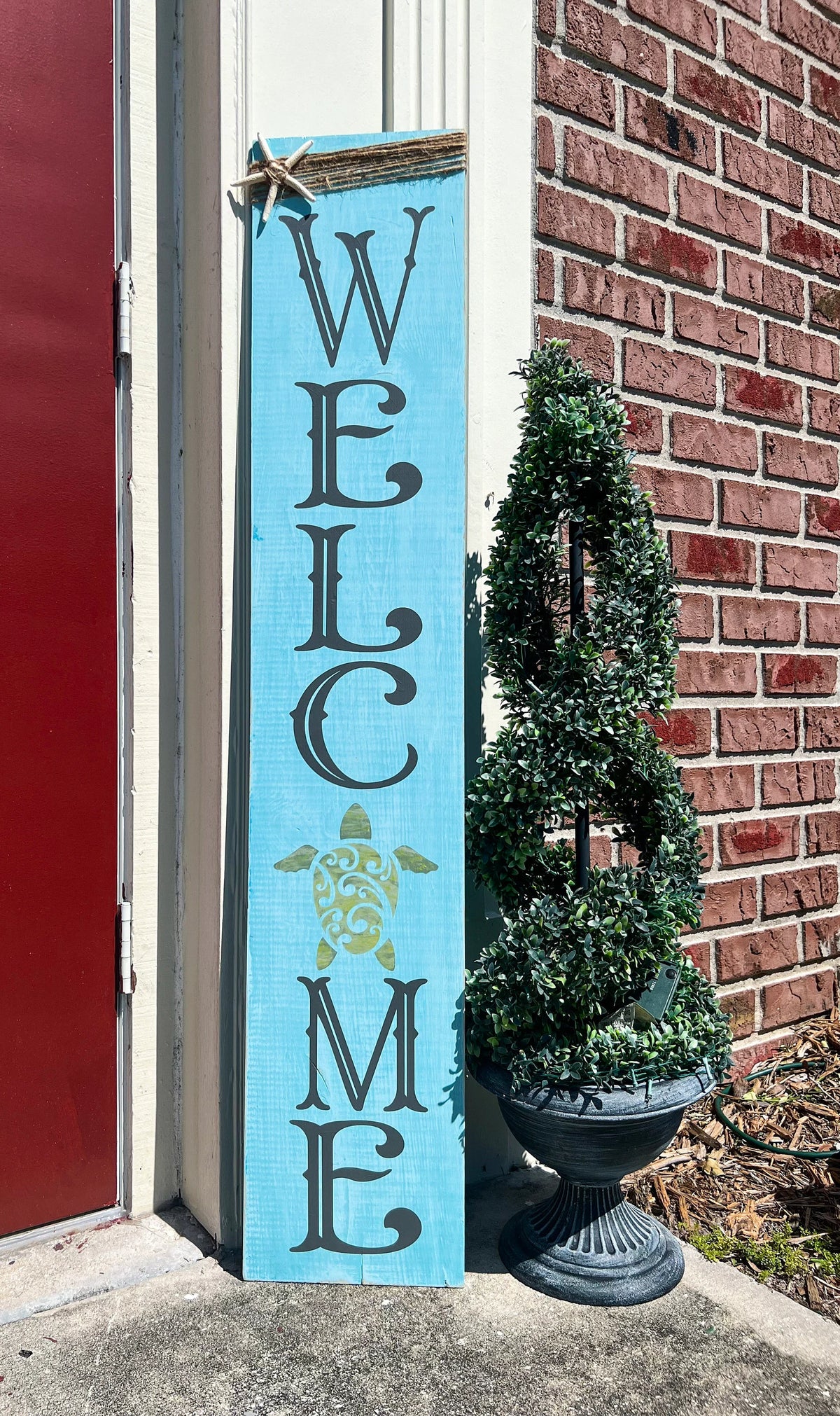 Sea turtle welcome aqua blue porch leaner sign with jute wrap and starfish leaning against white door beside boxwood topiary angled view