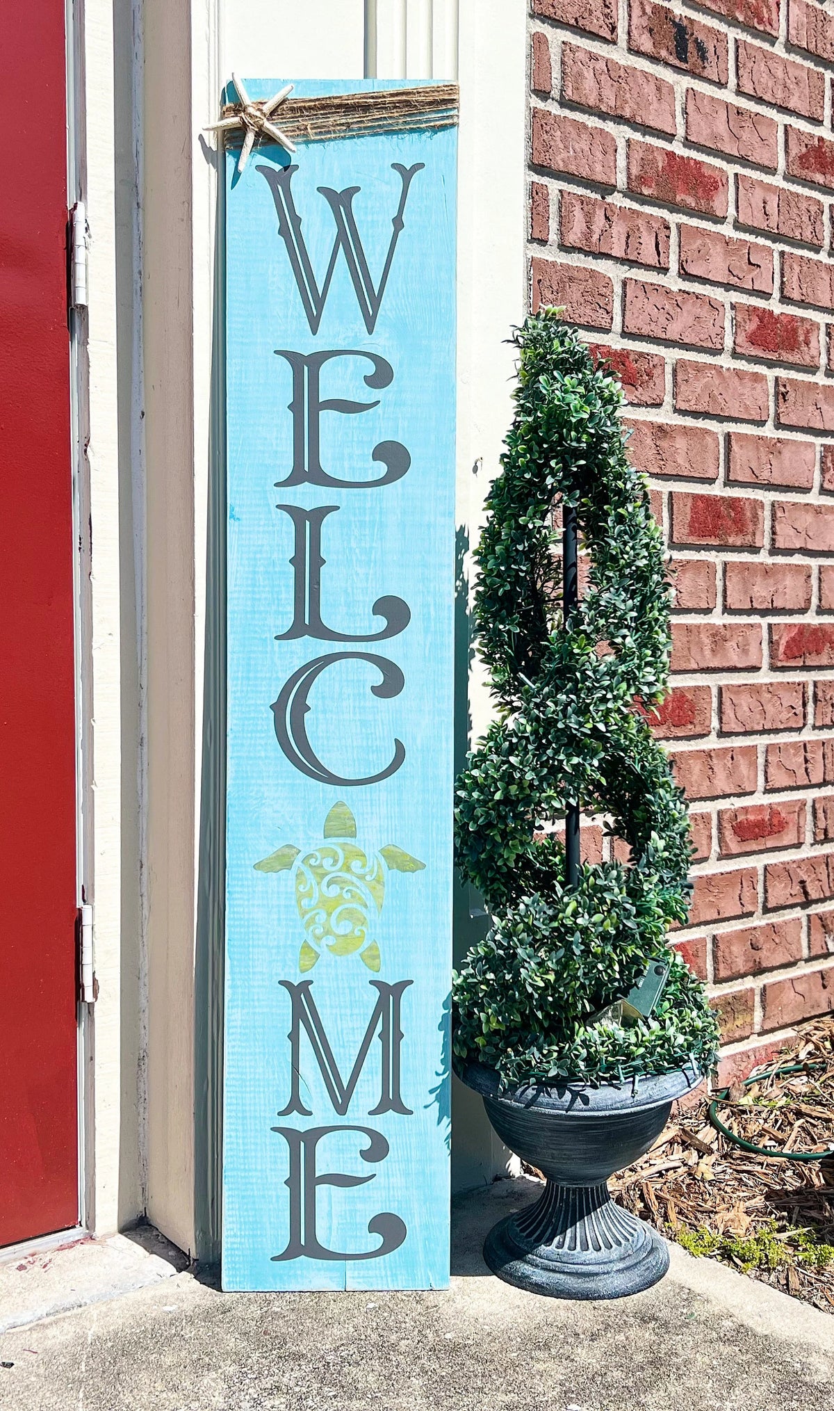 Sea turtle welcome aqua blue porch leaner sign with jute wrap and starfish leaning against white door beside boxwood topiary wide view
