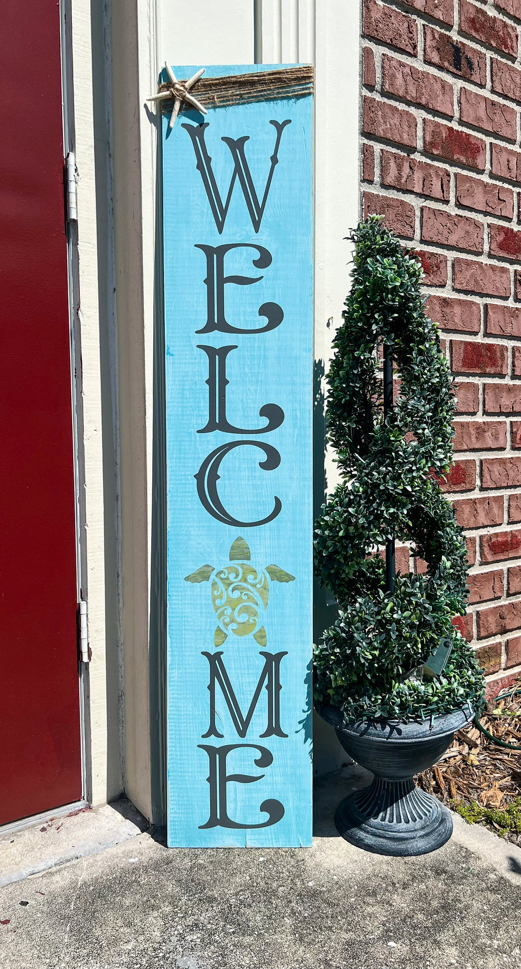 Sea turtle welcome aqua blue porch leaner sign with jute wrap and starfish leaning against white door beside boxwood topiary close up middle