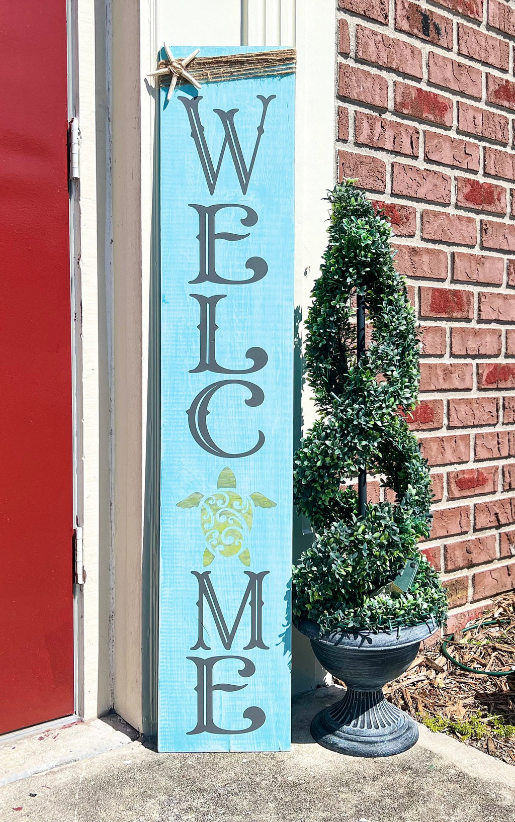 Sea turtle welcome aqua blue porch leaner sign with jute wrap and starfish leaning against white door beside boxwood topiary close up top
