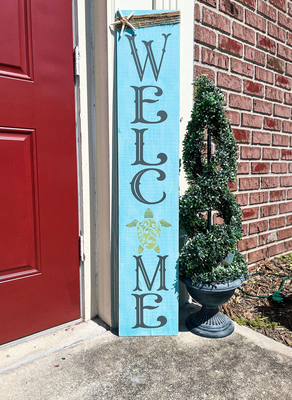 Sea turtle welcome aqua blue porch leaner sign with jute wrap and starfish leaning against white door beside red door and brick wall full view
