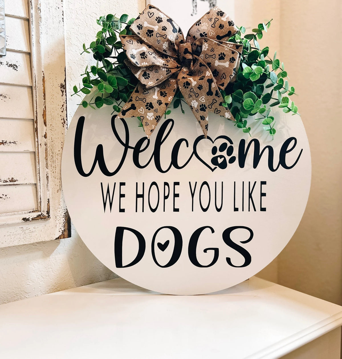 Welcome We Hope You Like Dogs all-white round pine door hanger with black lettering paw print heart O faux eucalyptus greenery and paw print burlap bow white colorway