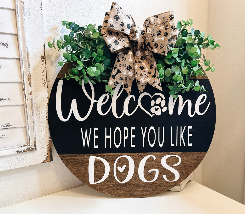 Welcome We Hope You Like Dogs two-tone round pine door hanger black upper half close up showing Welcome script with paw print heart O and WE HOPE YOU LIKE lettering with paw print burlap bow detail