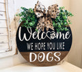 Welcome We Hope You Like Dogs two-tone round pine door hanger black upper half close up showing Welcome script with paw print heart O and WE HOPE YOU LIKE lettering with paw print burlap bow detail