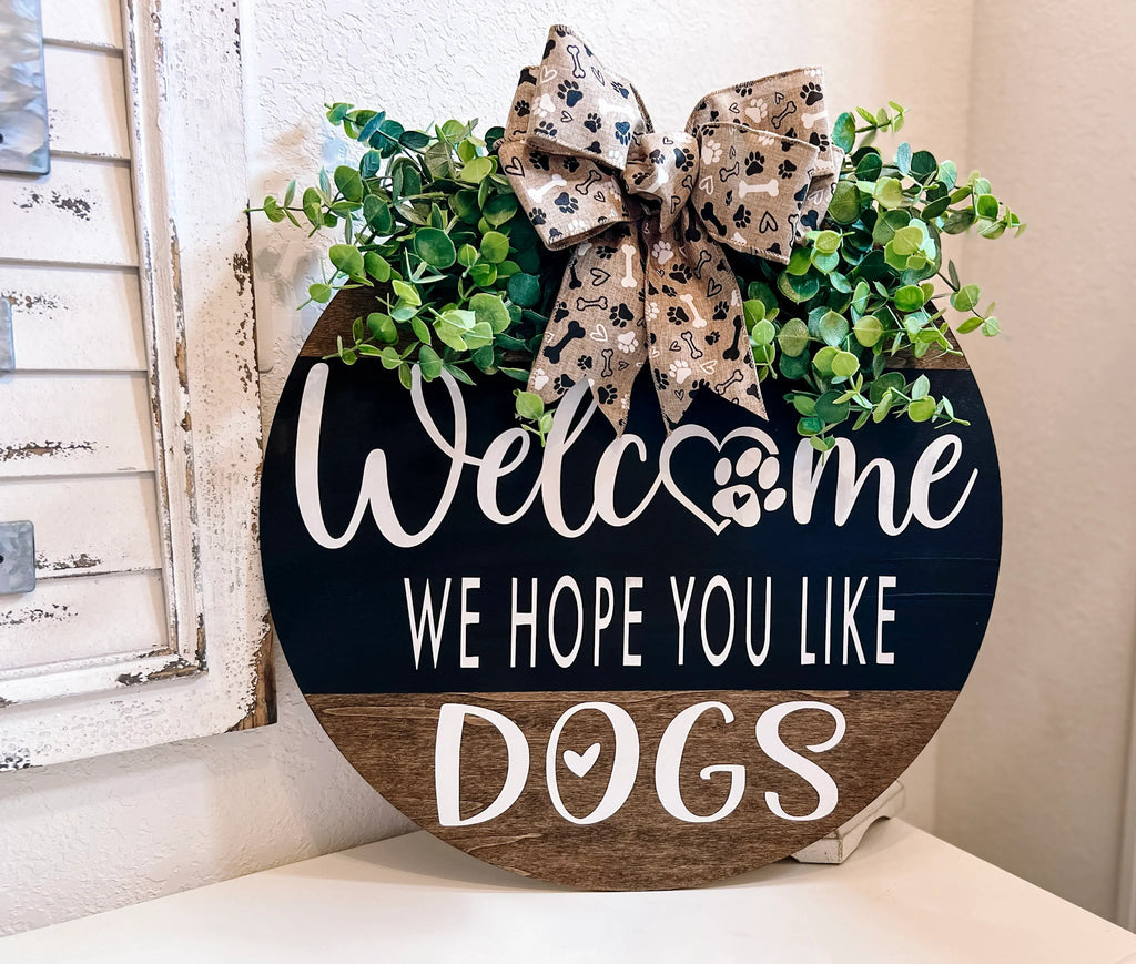 Welcome We Hope You Like Dogs two-tone round pine door hanger black upper half with white script and block lettering dark walnut lower half with DOGS lettering faux eucalyptus and paw print burlap bow on white surface front view