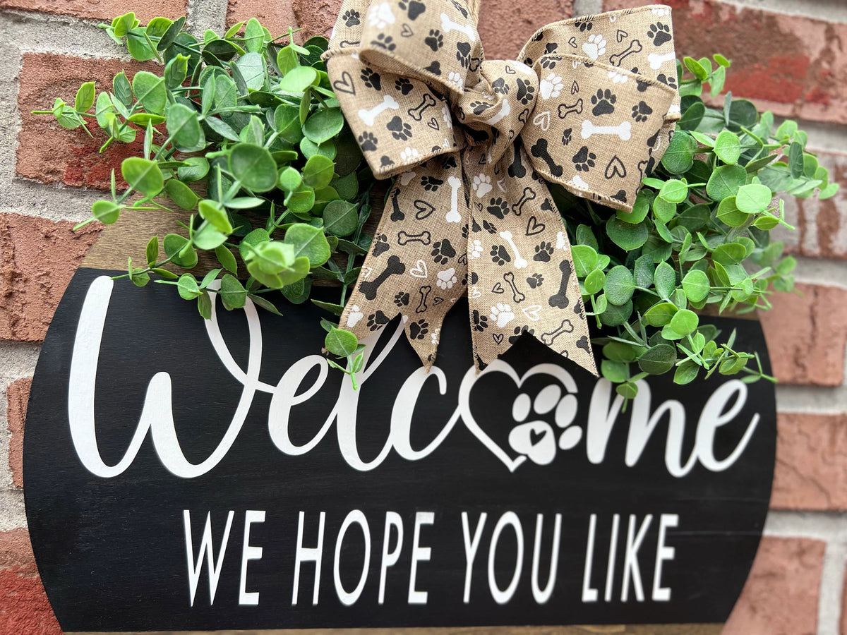 Welcome We Hope You Like Dogs two-tone round pine door hanger black upper half with white script and block lettering dark walnut lower half with DOGS lettering faux eucalyptus and paw print burlap bow on white surface angled view