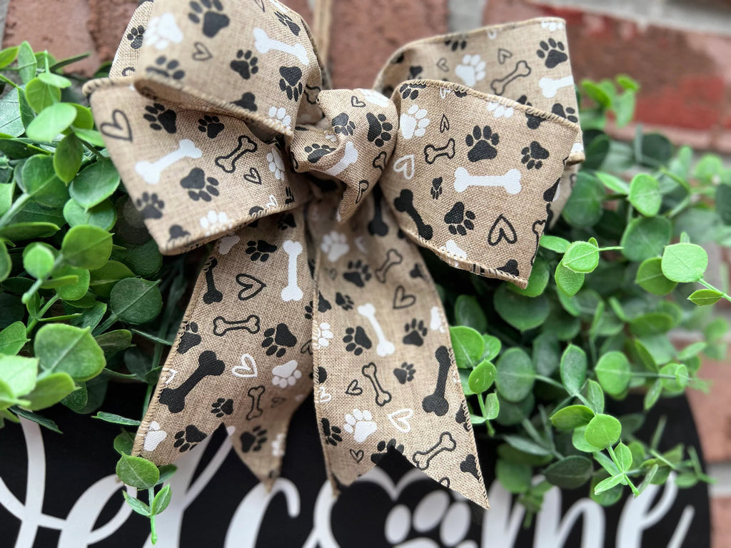 Welcome We Hope You Like Dogs two-tone round pine door hanger black upper half with white script and block lettering dark walnut lower half with DOGS lettering faux eucalyptus and paw print burlap bow leaning against white shutter wide shot