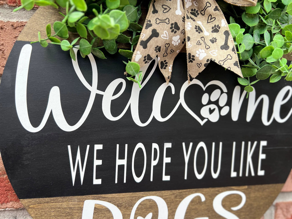 Welcome We Hope You Like Dogs two-tone round pine door hanger black upper half with white script and block lettering dark walnut lower half with DOGS lettering faux eucalyptus and paw print burlap bow leaning against white shutter hero shot